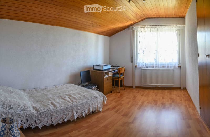 Cozy bedroom with wooden flooring, slanted wooden ceiling, and large window with sheer curtains. Features a neatly made bed, a wooden desk with a computer and printer, and a chair.