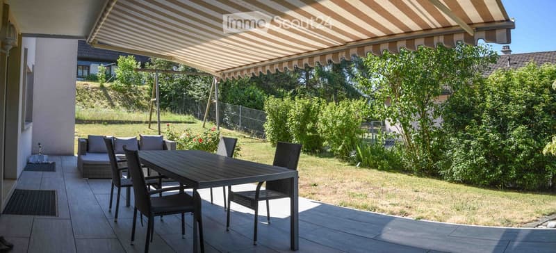 A covered patio area with a table, chairs, and a small sofa, surrounded by a well-maintained garden with trees and shrubs.