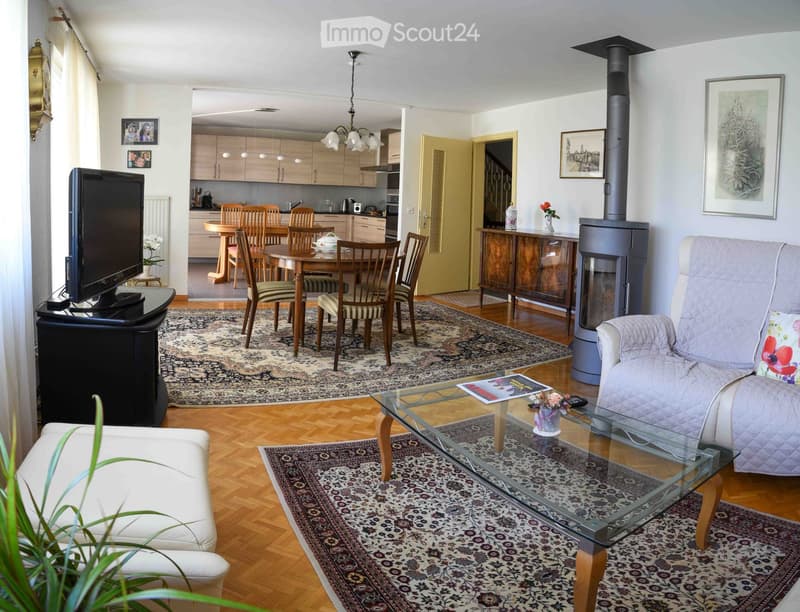 Spacious living room with hardwood floors, a large sofa, a glass coffee table, a television, a dining area with a table and chairs, and a fireplace. The room is decorated with rugs, framed pictures, and a vase with flowers.