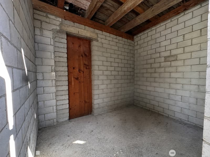 Empty room with exposed brick walls, wooden door, and wooden ceiling beams. Concrete floor, no furniture or equipment.