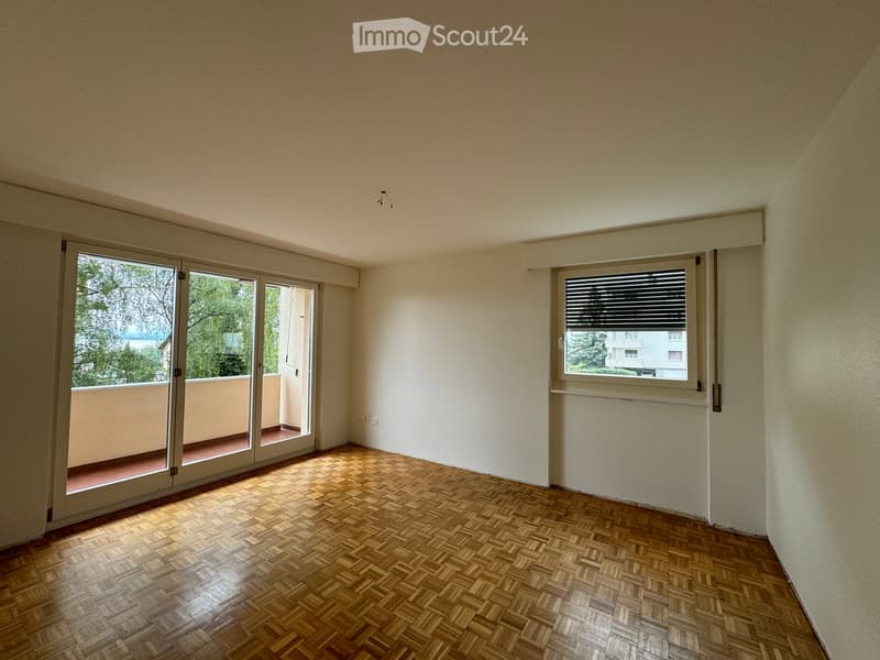 Empty room with wooden parquet flooring, white walls, large sliding glass doors leading to a balcony, and a window with blinds.