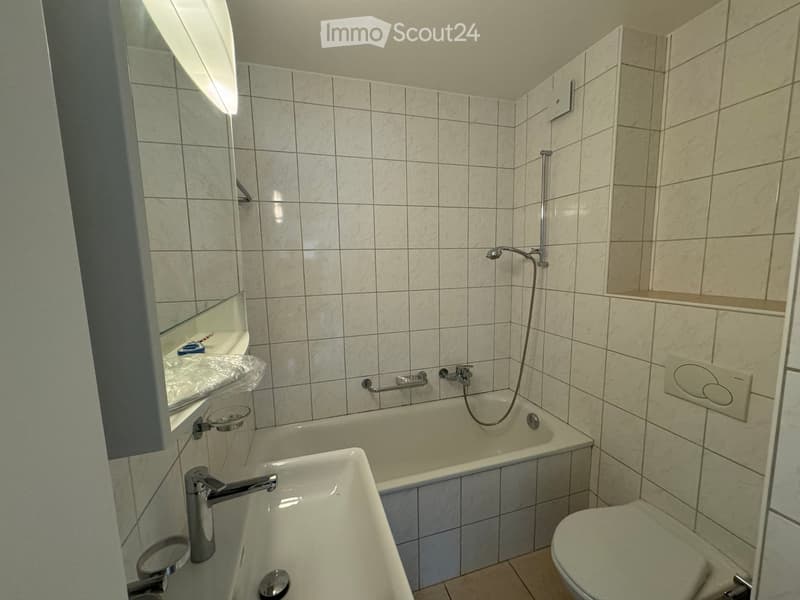 Bathroom with tiled walls and floor, featuring a bathtub, sink, toilet, and a mirror with a built-in shelf. Includes a handrail for safety.