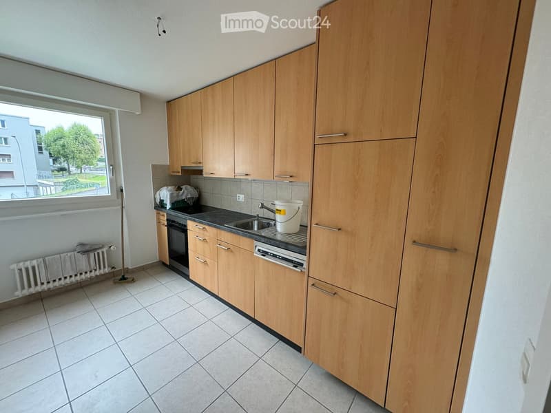Modern kitchen with light wooden cabinets, tiled floor, and stainless steel appliances including a sink, oven, and dishwasher. A white bucket is placed on the counter.