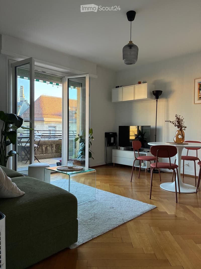 Modern living room with wooden flooring, a green sofa, a glass coffee table, a white dining table with red chairs, a television, and a balcony with outdoor furniture.