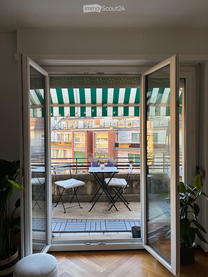 Balcony with a small table, two chairs, a stool, and potted plants, overlooking a cityscape with striped awnings.