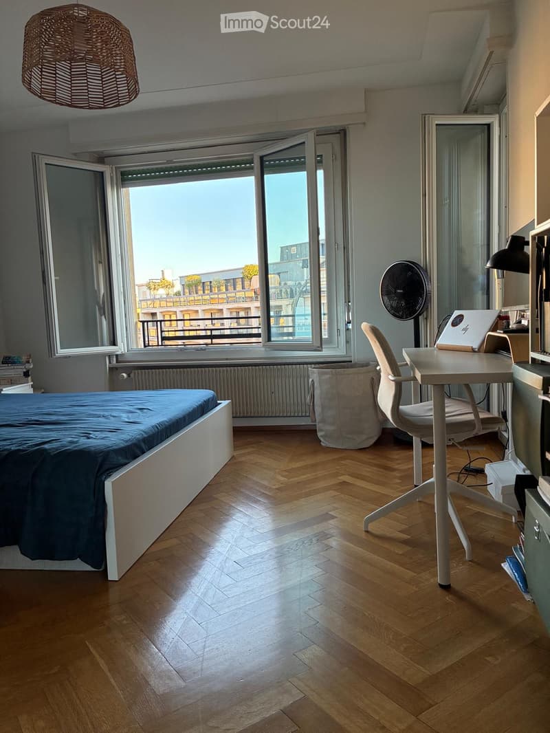 Bright bedroom with wooden flooring, large windows offering a city view, a bed with blue bedding, a desk with a laptop, a chair, a fan, and a wicker basket.