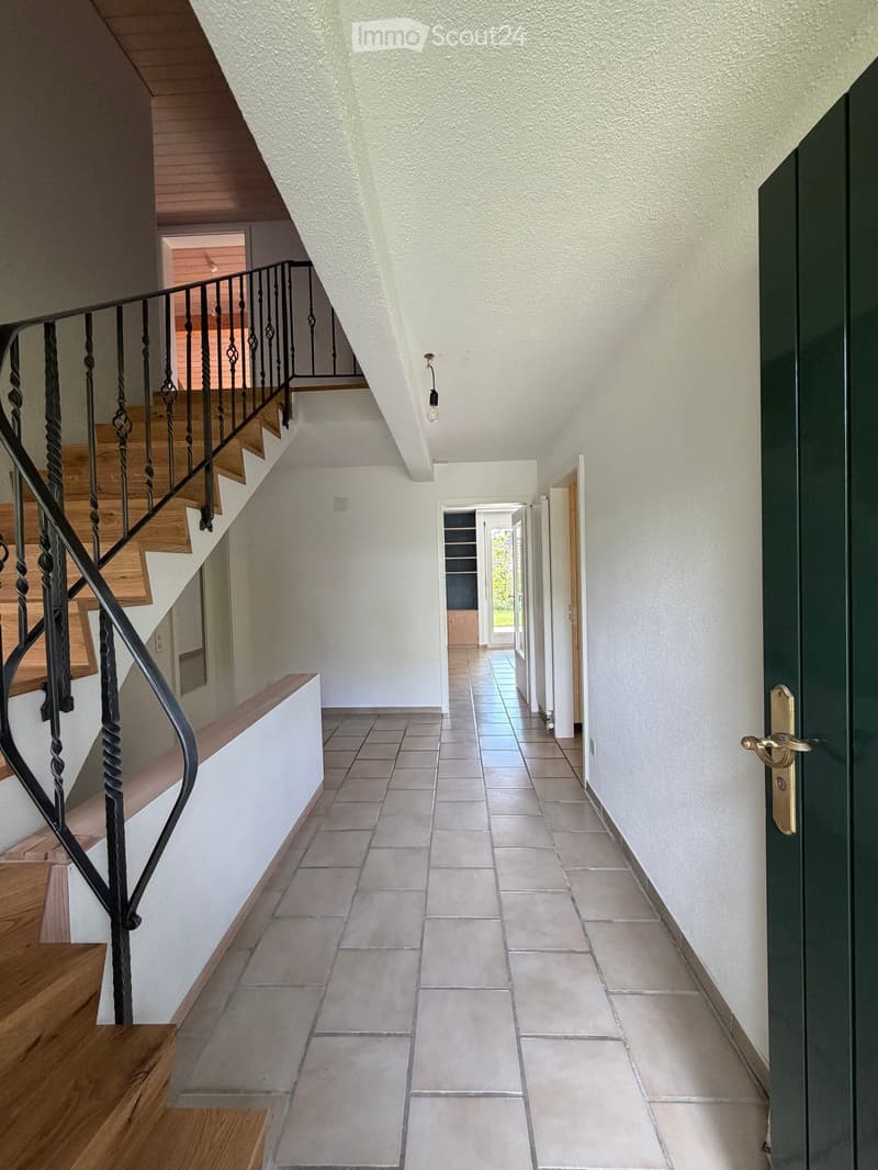 Spacious hallway with tiled floor, white walls, wooden staircase with black wrought-iron railing, and a green door at the end.