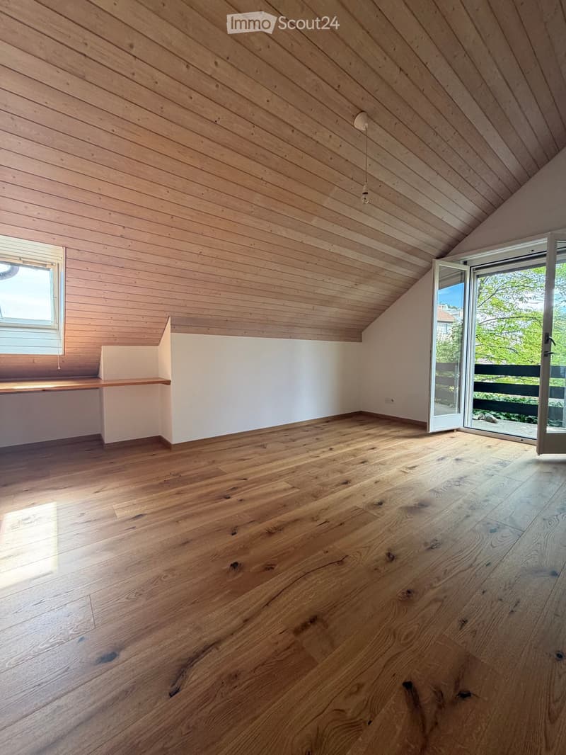 Completely empty room with wooden floor and slanted wooden ceiling, featuring a window and an open sliding door leading to a balcony.