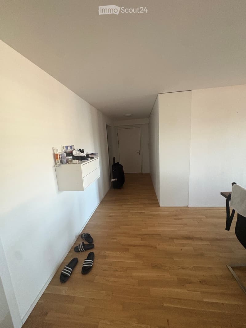 A narrow hallway with light wooden flooring and white walls, featuring a small shelf with personal items, a suitcase, and a pair of slippers on the floor.