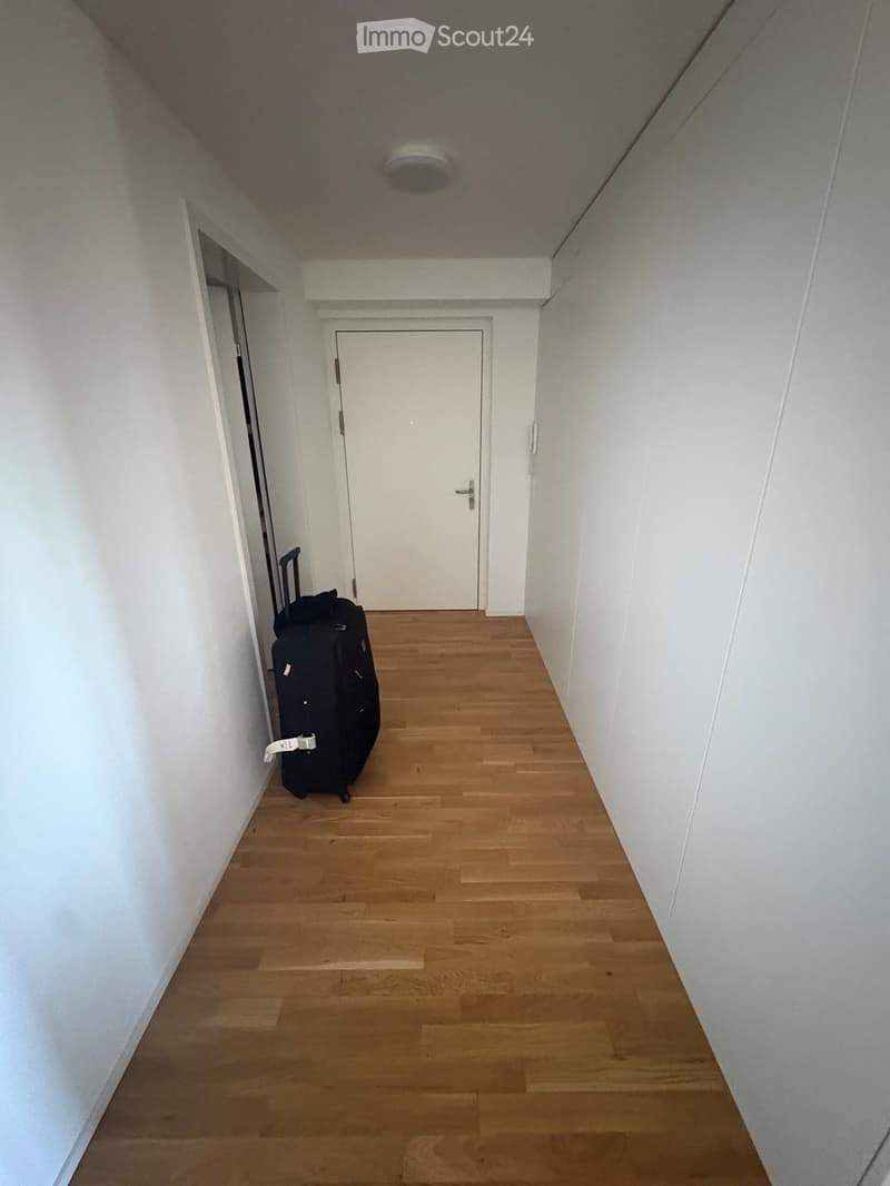 Empty hallway with wooden flooring, white walls, and a closed door at the end. A single black suitcase is placed against the left wall.