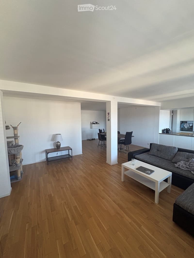 Modern living room with wooden flooring, white walls, and a grey sofa. Features a white coffee table, a small table lamp, a cat tree, and a dining area visible in the background.