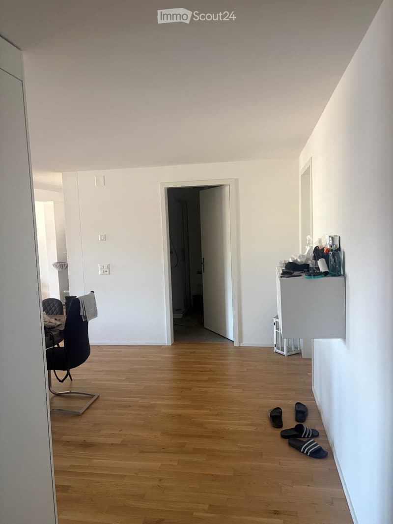 A modern, minimalistic room with light wooden flooring, white walls, and a large open doorway leading to another space. A black chair and a small table are positioned near the left wall, while a mirror and a shelf with various items are on the right side.
