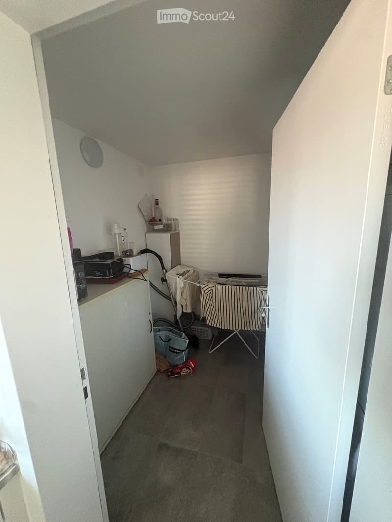 A narrow hallway with a tiled floor, white walls, and a small closet with shelves containing various items. A partially visible ironing board and some scattered items are present.