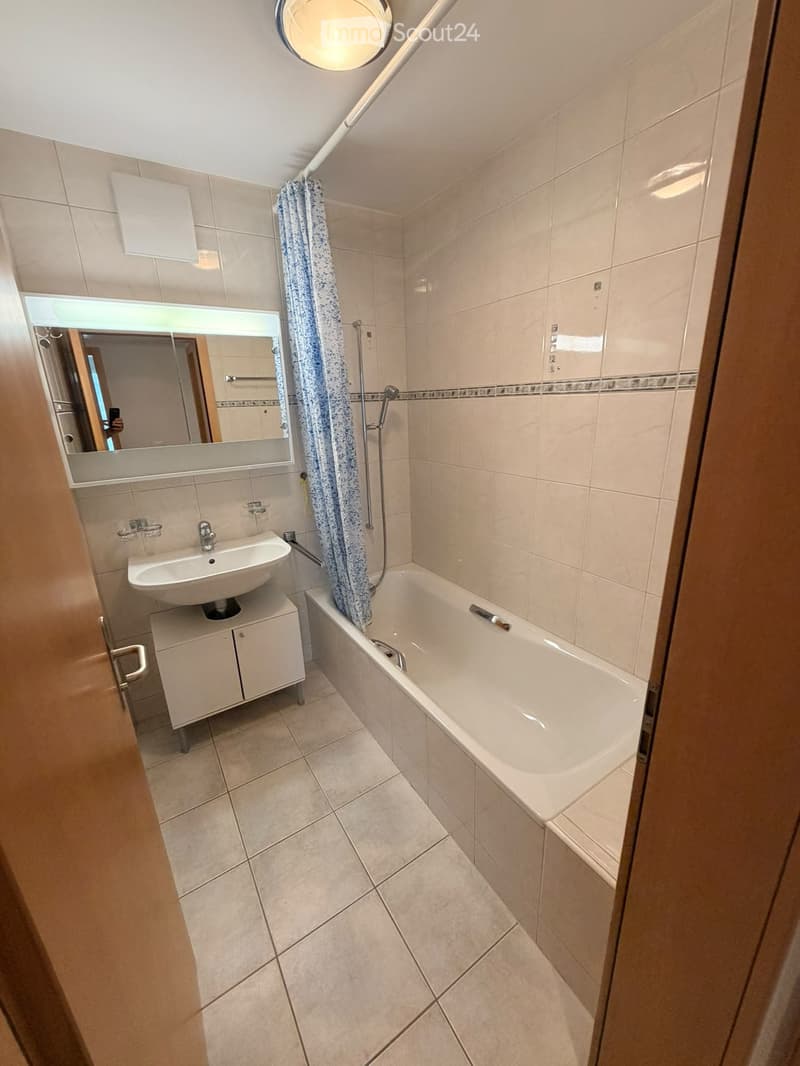 Modern bathroom with a bathtub, sink, mirror, and cabinet. Features tiled walls and floor, a shower curtain, and a round ceiling light.