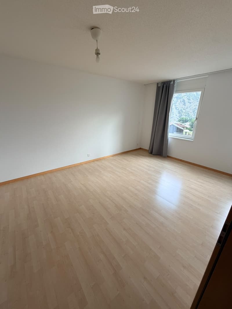 Empty room with light wooden flooring, white walls, and a single window with dark curtains. A ceiling light fixture is visible.