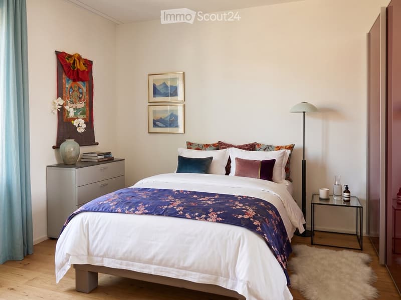 A well-furnished bedroom with a large bed, nightstand, floor lamp, decorative vase, books, and a small table with a cup and bottle. The room features a wooden floor and light-colored walls.