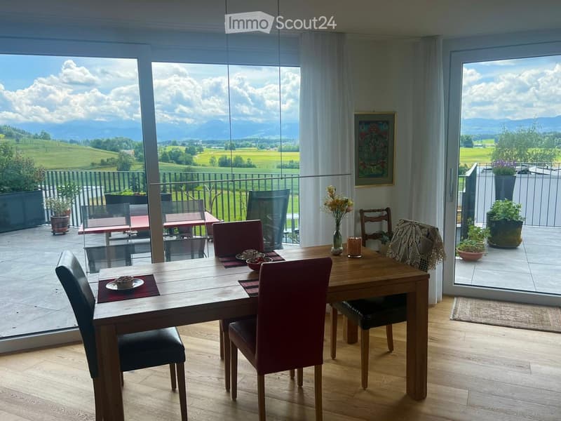 Bright dining room with wooden floor, large windows offering scenic mountain views, dining table with chairs, vase with flowers, and decorative items.