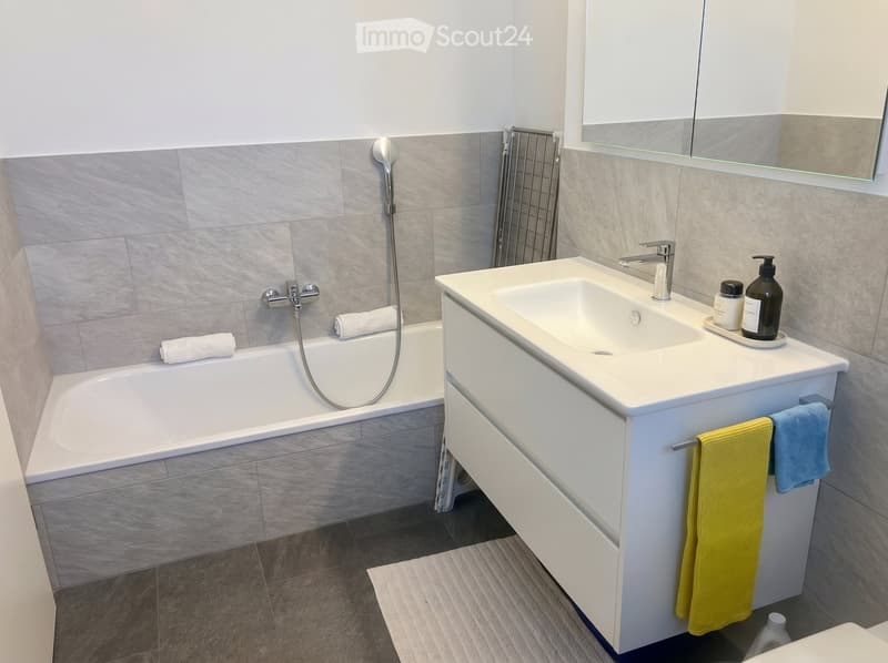 Modern bathroom with a bathtub, sink, and mirror. Includes towels and toiletries on the sink counter.