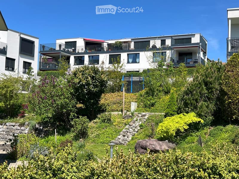 Modern multi-story residential building with balconies, surrounded by lush greenery and a landscaped garden area.
