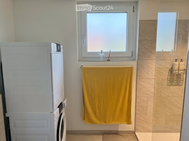 Bathroom with a washing machine, a yellow towel hanging on a rack, a toothbrush, and a soap dispenser on the windowsill.