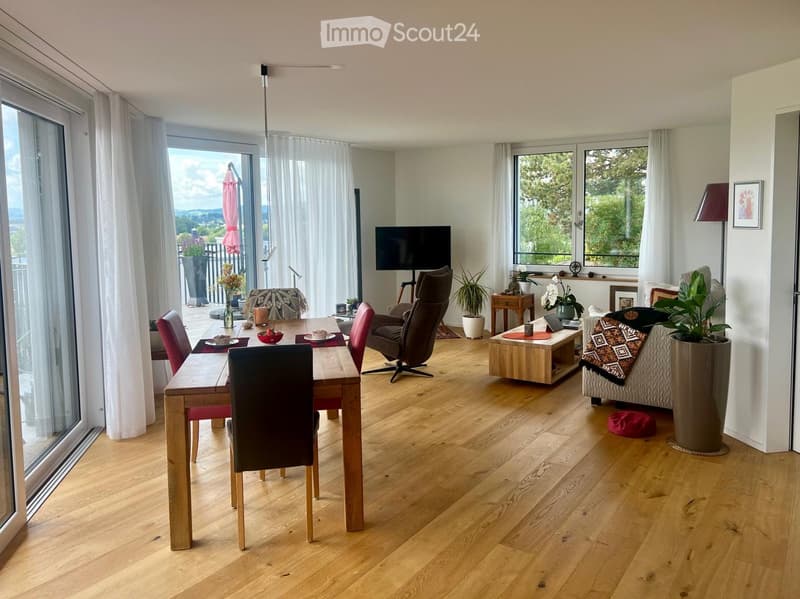 Open-plan living and dining area with wooden flooring, large windows offering a scenic view, a sofa, coffee table, dining table with chairs, television, and decorative plants.