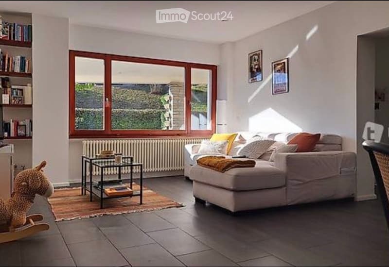 Bright living room with large windows offering a scenic view, featuring a light-colored sofa with colorful cushions, a small coffee table, a rocking horse, and a bookshelf with books and decorative items.