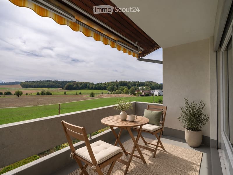 Balcony with a wooden table, two chairs, a cup, a vase with flowers, and a potted plant, overlooking a scenic countryside view.