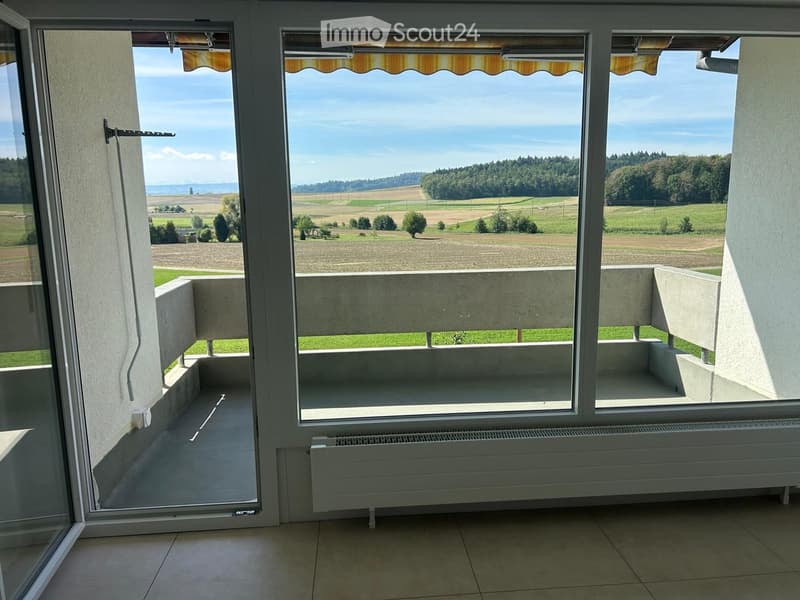 Balcony with large windows offering a scenic view of fields and trees, featuring built-in seating and a radiator below the windows.