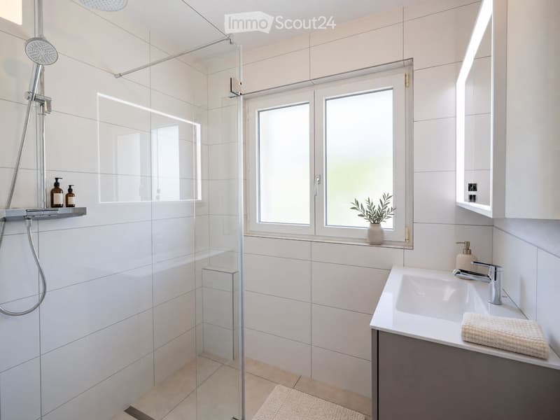 Modern bathroom with white tiles, glass shower enclosure, double sink, and a small plant on the windowsill.