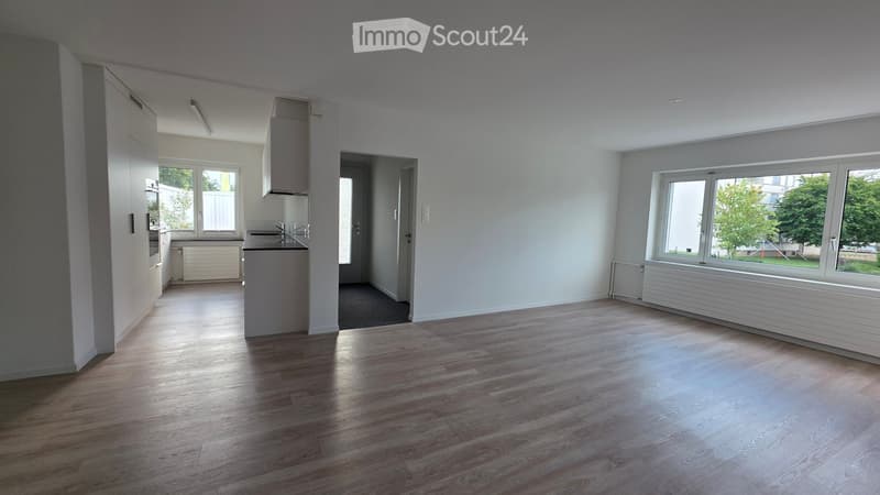 Empty room with wooden flooring, white walls, large windows, and a small kitchen area visible in the background.