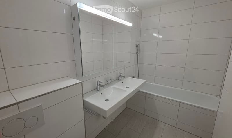 Modern bathroom with a double sink, bathtub, large mirror with built-in lighting, and sleek cabinetry.