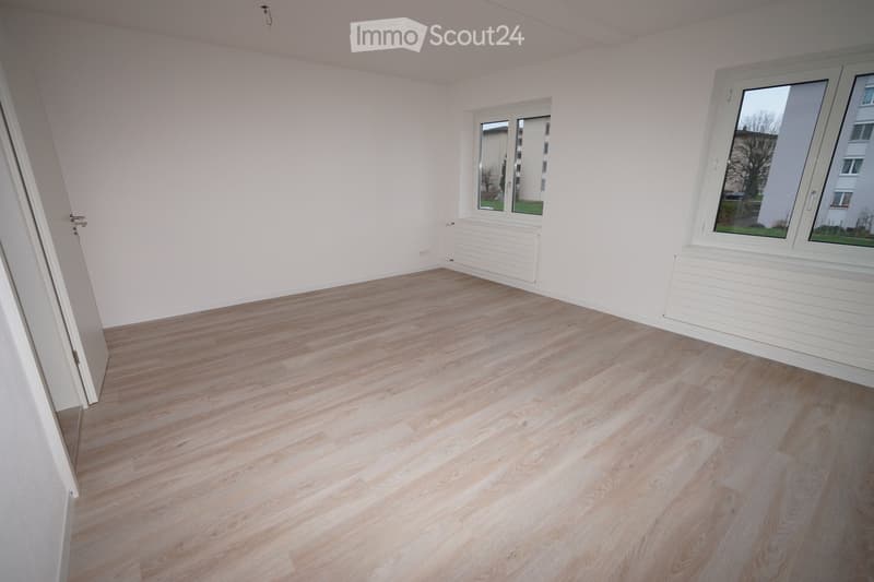 Empty room with light wooden flooring and white walls, featuring two windows and a door.