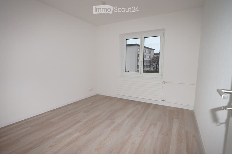 Empty room with light wooden flooring, white walls, and a large window with white frame. The room has a radiator below the window and a door visible on the right side.