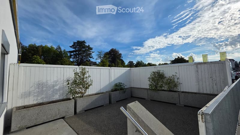 Balcony with white wooden fence, concrete planters with small plants, metal handrail, and a clear blue sky with scattered clouds.