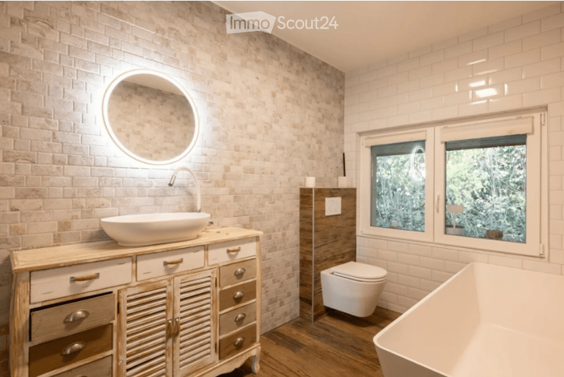 Modern bathroom with a wooden vanity cabinet, round illuminated mirror, white sink, toilet, and freestanding bathtub. The walls are tiled in a light, textured pattern, and the floor is wooden.
