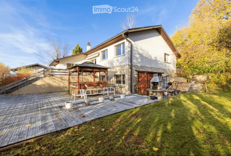 A modern two-story house with a wooden deck, outdoor seating area, and a gazebo. The garden features a well-maintained lawn, potted plants, and a barbecue area.