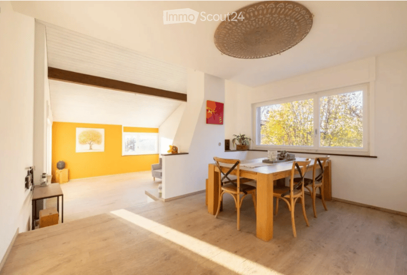 Bright, open-plan dining area with light wooden flooring, a white dining table surrounded by chairs, a potted plant on the windowsill, and artwork on the walls.