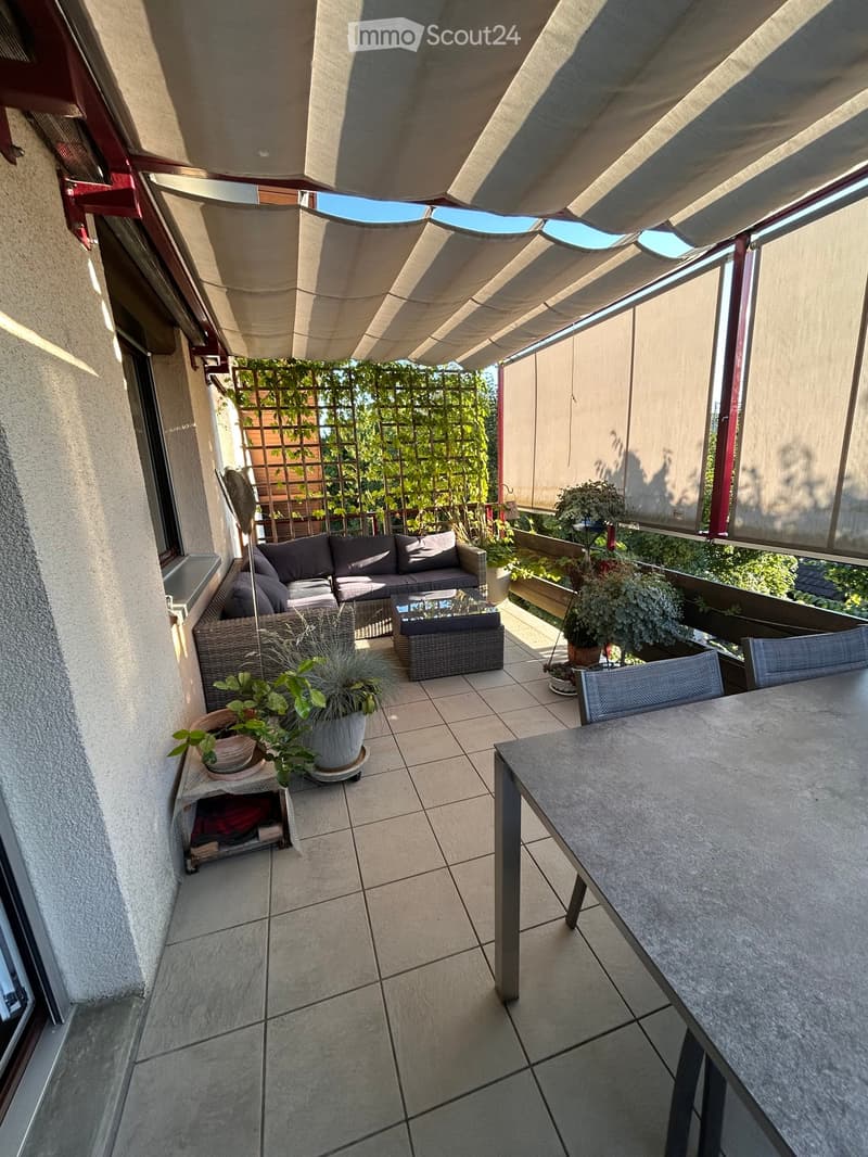 Balcony with tiled floor, covered by a retractable awning, featuring a grey sofa, coffee table, dining table, chairs, and potted plants.