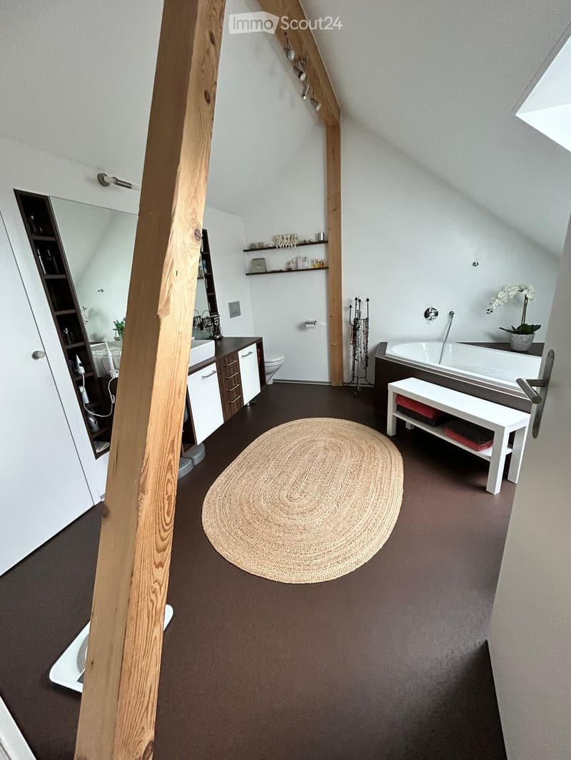 Modern bathroom with dark flooring, white walls, a round woven rug, a white bench, a bathtub, a sink with cabinet, and a mirror. Shelves hold decorative items and toiletries.