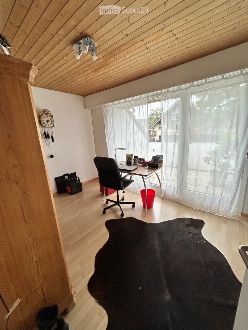 Bright, slanted-ceiling room with wooden flooring, a desk with a chair, a laptop, a clock, a lamp, and a large black rug.