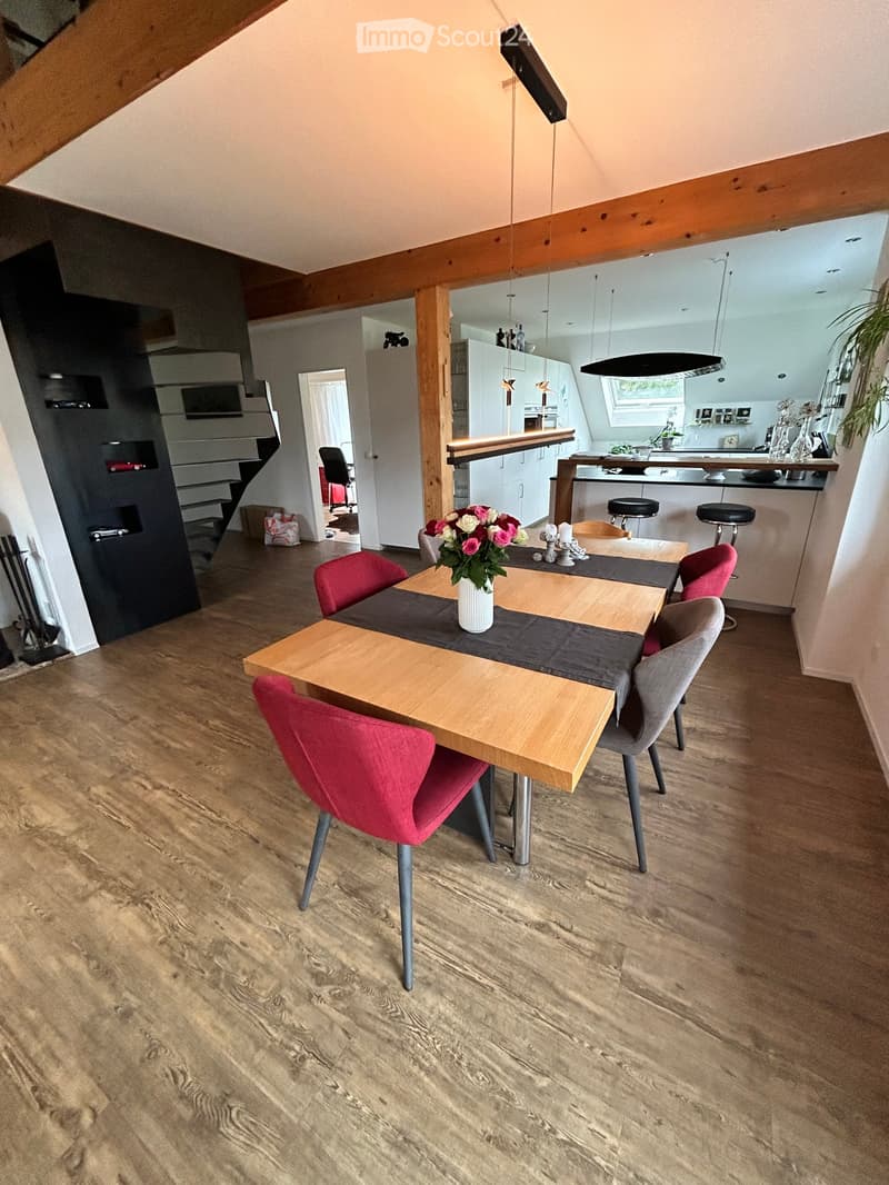 Dining area with wooden table, red chairs, grey chairs, and a vase with flowers. Open kitchen area visible in the background with white cabinets and a kitchen island.