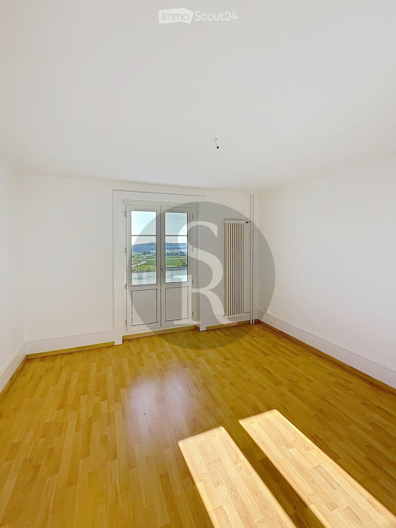 Empty room with light wooden flooring, white walls, a large window with a view, and a radiator on one wall.