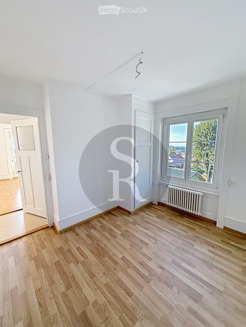 Empty room with light wooden flooring, white walls, and large windows offering a view of the outside. A radiator is visible under one window, and a door leads to another room.