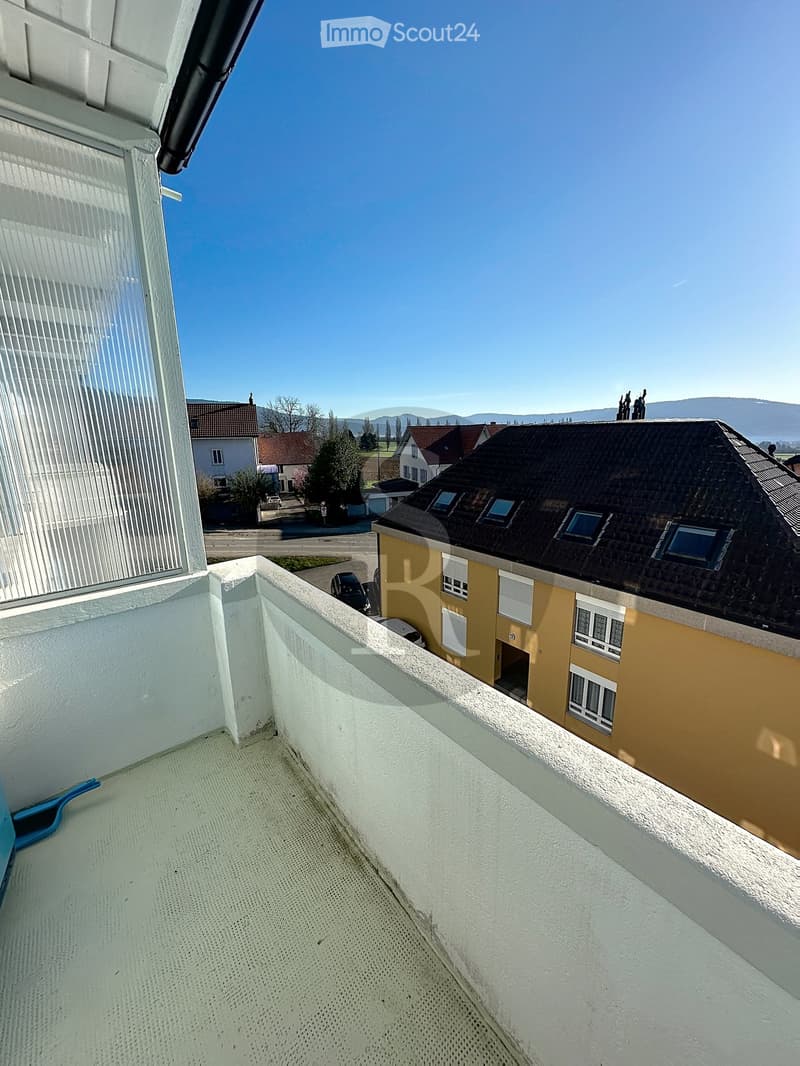 Balcony with white walls and a metal safety railing, overlooking a residential area with houses, trees, and a clear blue sky.
