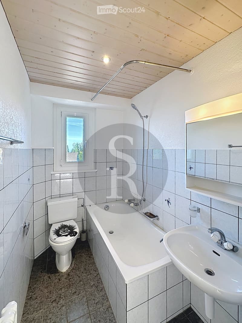 Modern bathroom with white tiled walls, a bathtub, a toilet, a sink, a mirror, and a showerhead. The room features a window with a view and a built-in shelf above the sink.