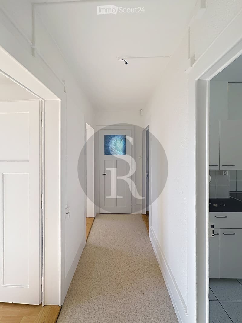 A clean, well-lit hallway with white walls, carpeted floor, and several doors leading to different rooms. The hallway features a central door with a frosted glass panel and a side door leading to a kitchen area with white cabinets and a tiled floor.