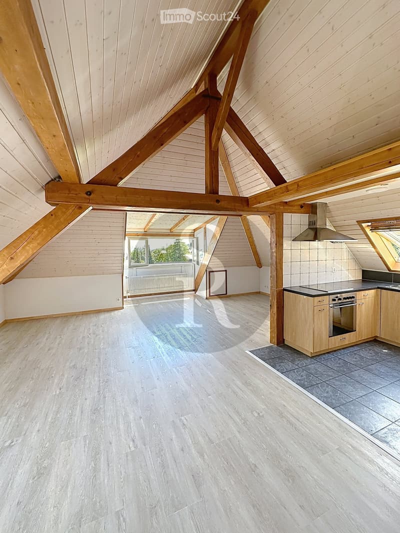 Open-plan attic space with wooden beams, light wooden flooring, and a small kitchen area featuring cabinets, an oven, and a range hood. A large window offers a view of greenery outside.