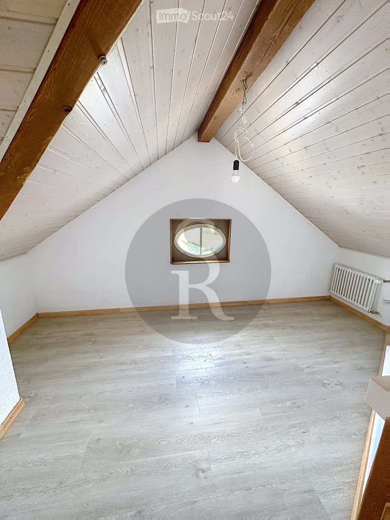 An empty attic room with wooden flooring, white walls, and exposed wooden beams on the ceiling. Features a skylight window and a radiator on one wall.