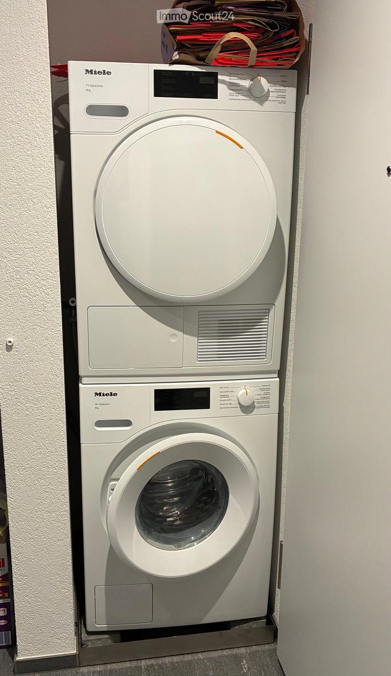 White Miele washing machine and dryer stacked vertically in a bathroom, with control panels and a small storage box on top.