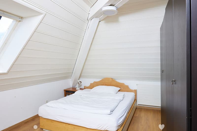 Cozy attic bedroom with wooden bed, nightstand, and lamp. Slanted white paneled ceiling and large windows provide natural light.